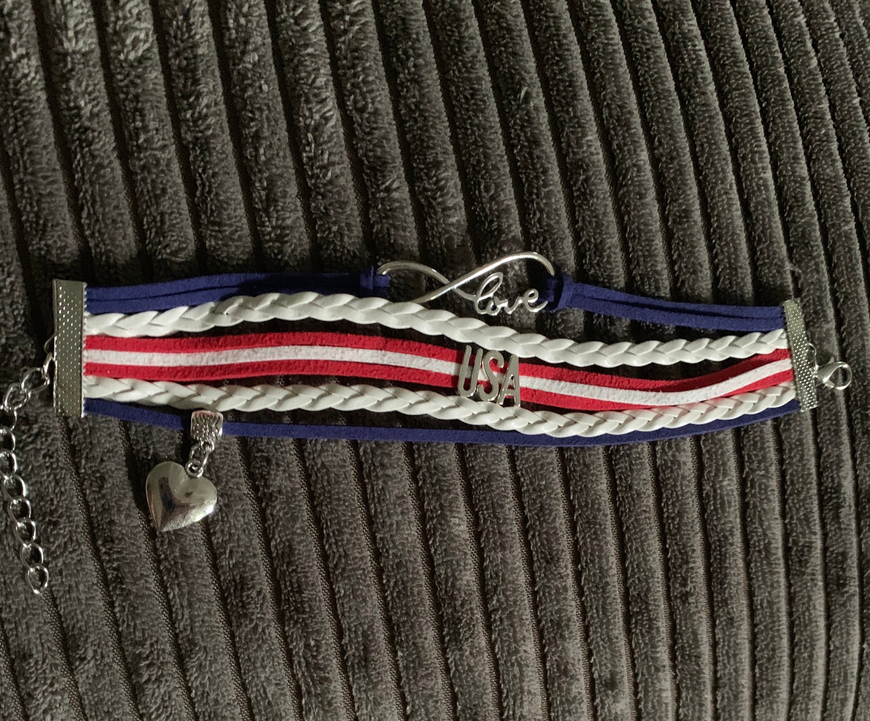 USA patriotic bracelet made with 5 bands connected with silver clasps to make a one piece bracelet. Bands contain braided and straight strings or ribbon adorned with metal attachments like USA lettering and a dangling silver heart. Very nice quality bracelet.  Clasp chain can adjust for sizing. 